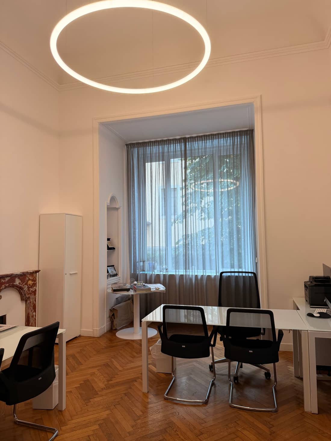 La sala riunioni dello studio: tavolo bianco con due sedie di fronte, finestra alta con tenda chiara e lampada ad anello